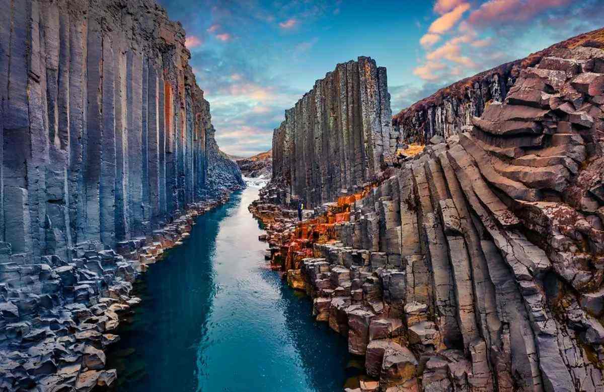 A stunning view of a river cutting through dramatic basalt columns in Iceland. The towering, geometric rock formations are reflected in the calm blue water, while the sky is lit up with warm colors from the setting sun.