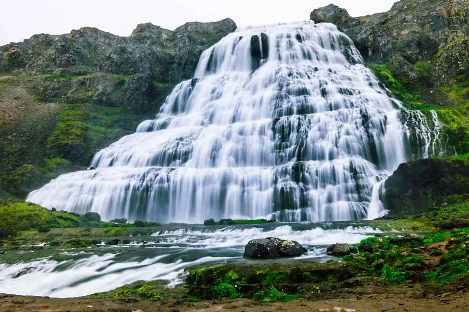 Close-up view of Dynjandi Waterfall, a tiered waterfall located in the Westfjords of Iceland, cascading down a rocky mountain. The surrounding green moss and rugged cliffs enhance the beauty of the flowing water.