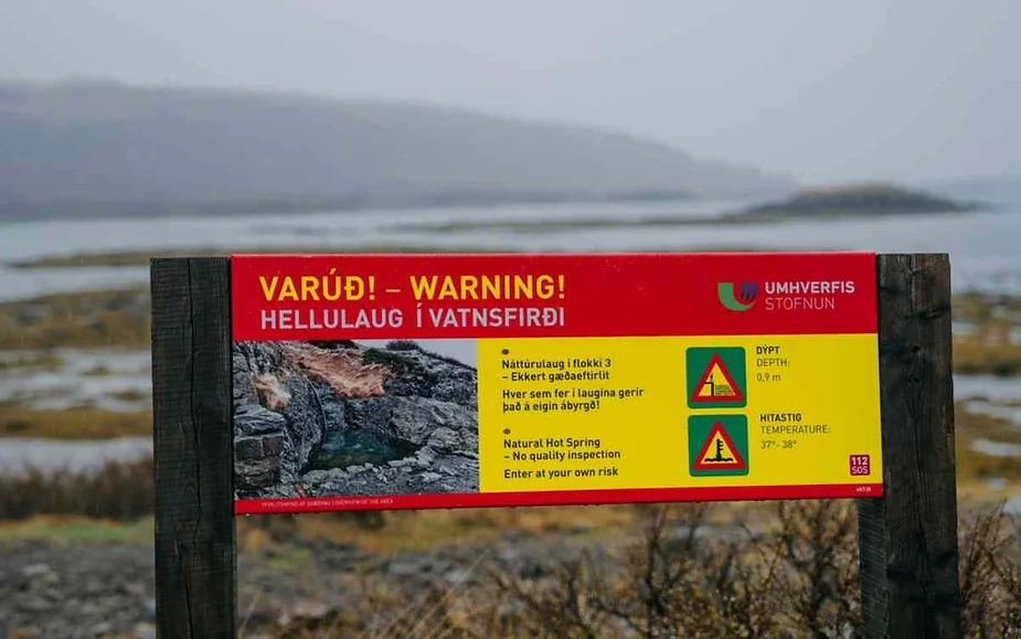 A warning sign at Hellulaug natural hot spring in Vatnsfjörður, Iceland. The sign, written in both Icelandic and English, cautions visitors about the lack of quality inspections and advises them to enter at their own risk. The background shows a misty coastal landscape with rocks and water.