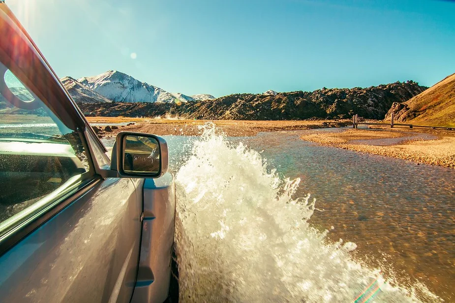 Close-up of a vehicle driving through a shallow river with a scenic Icelandic landscape, including mountains and blue skies in the background