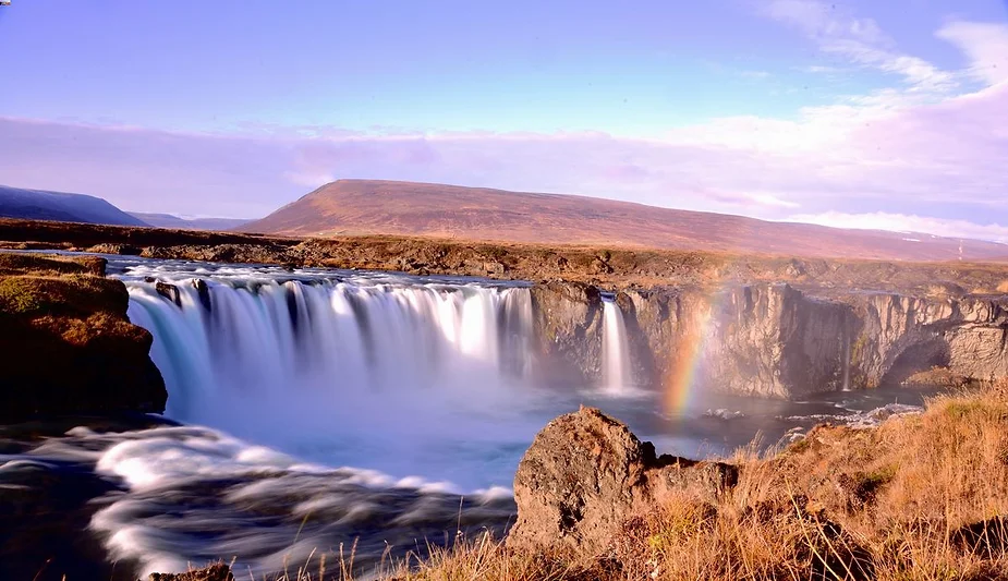 Scenic view of Godafoss waterfall in Iceland, with a rainbow forming in the mist, surrounded by rugged cliffs and a clear blue sky