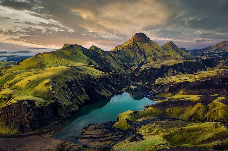 Stunning aerial view of Iceland's highlands featuring rugged green mountains, a tranquil turquoise lake, and dramatic clouds during sunset