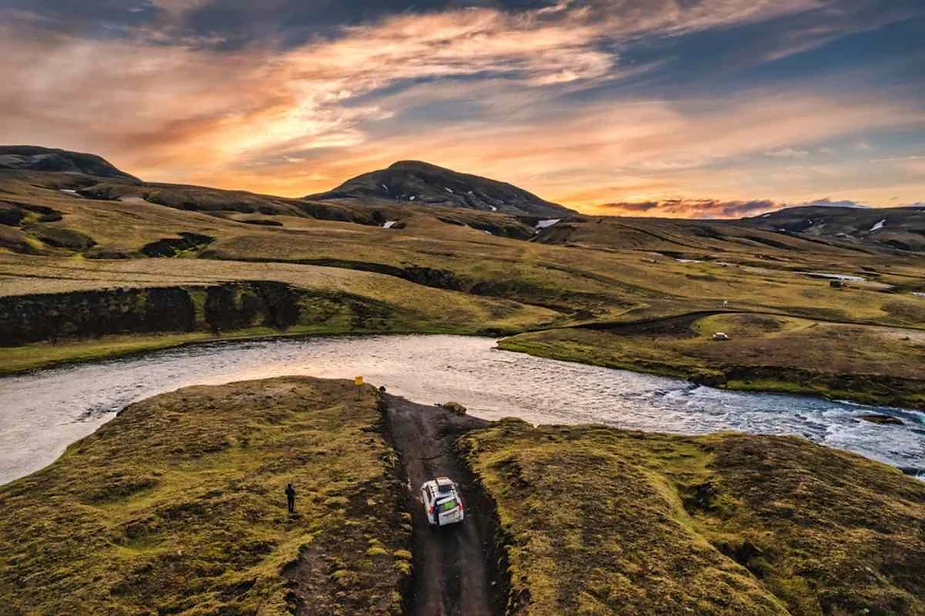 A 4x4 vehicle navigates a dirt road through Iceland’s remote highlands at sunset, crossing a river amidst rolling hills and dramatic skies. The scene emphasizes the adventurous spirit of off-road travel in Iceland’s wilderness.