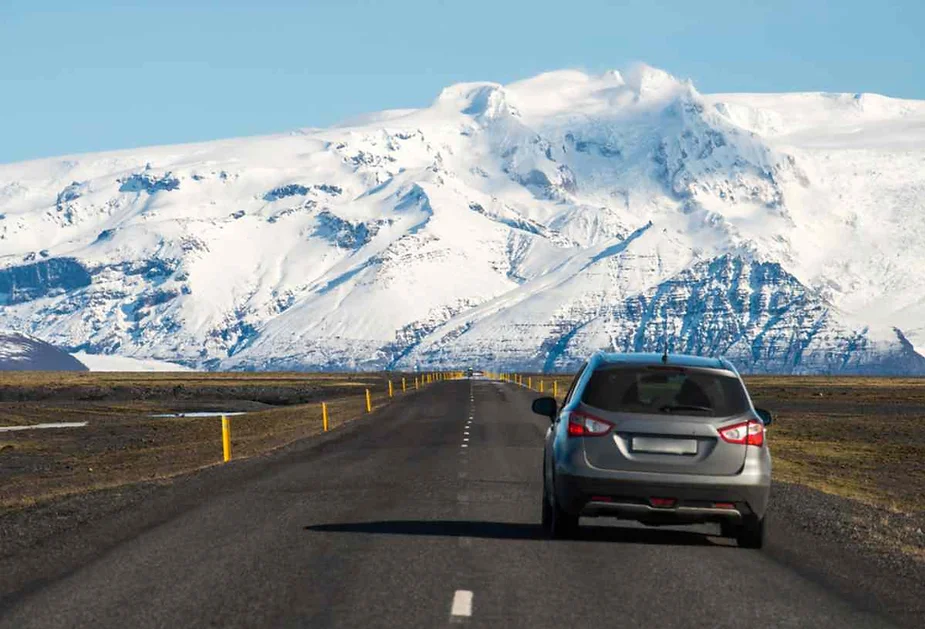 A silver car driving on a clear road toward Iceland's snow-covered mountains, showcasing the dramatic landscape and open road conditions ideal for a road trip across Iceland.