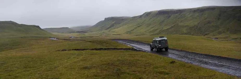 A rugged 4x4 vehicle drives along a remote gravel road through Iceland's vast, green highlands under cloudy skies. The image highlights off-road driving in Iceland's rugged terrain.