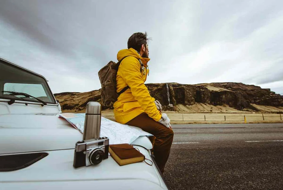 A traveler wearing a yellow jacket sits on the hood of a car parked on an Icelandic road, with a map, camera, and thermos beside them. The scenic landscape, featuring cliffs and a distant waterfall, emphasizes a road trip adventure through Iceland’s unique natural beauty.