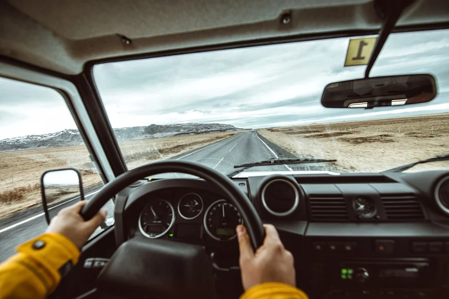 A driver navigates a straight, open road through Iceland's barren landscape, viewed from the vehicle's interior, with hands on the steering wheel