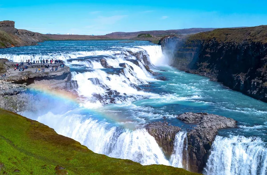 A large group of tourists admire the powerful Gullfoss waterfall in Iceland, with cascading water and a rainbow forming at the base of the falls