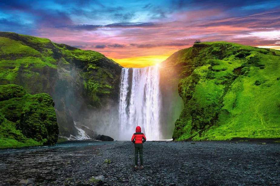 A person in a red jacket stands in front of the majestic Skógafoss waterfall in Iceland, with lush green cliffs and a colorful sunset in the background