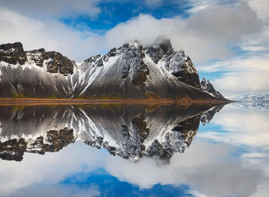 A stunning reflection of the snow-capped Vestrahorn mountain in Iceland, mirrored perfectly on a calm, clear lake under a partly cloudy sky