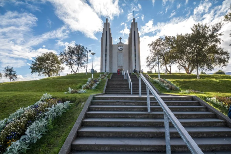 A view of the Church of Akureyri in Iceland, with its iconic twin towers, surrounded by lush greenery and a blue sky