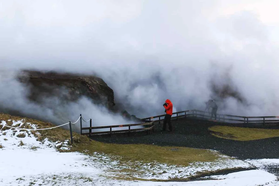 A tourist in a red jacket photographs a steaming geothermal area in Iceland, with snow-covered ground and rising steam creating a surreal and misty landscape.