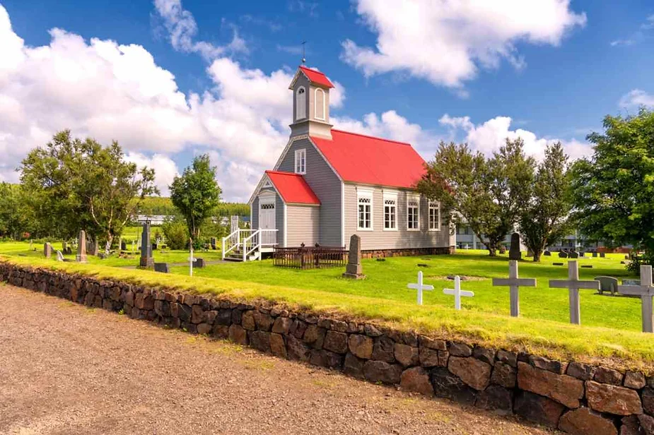 A charming white church with a red roof stands amidst a small cemetery in a peaceful Icelandic village, surrounded by greenery and framed by a stone wall under a bright blue sky.