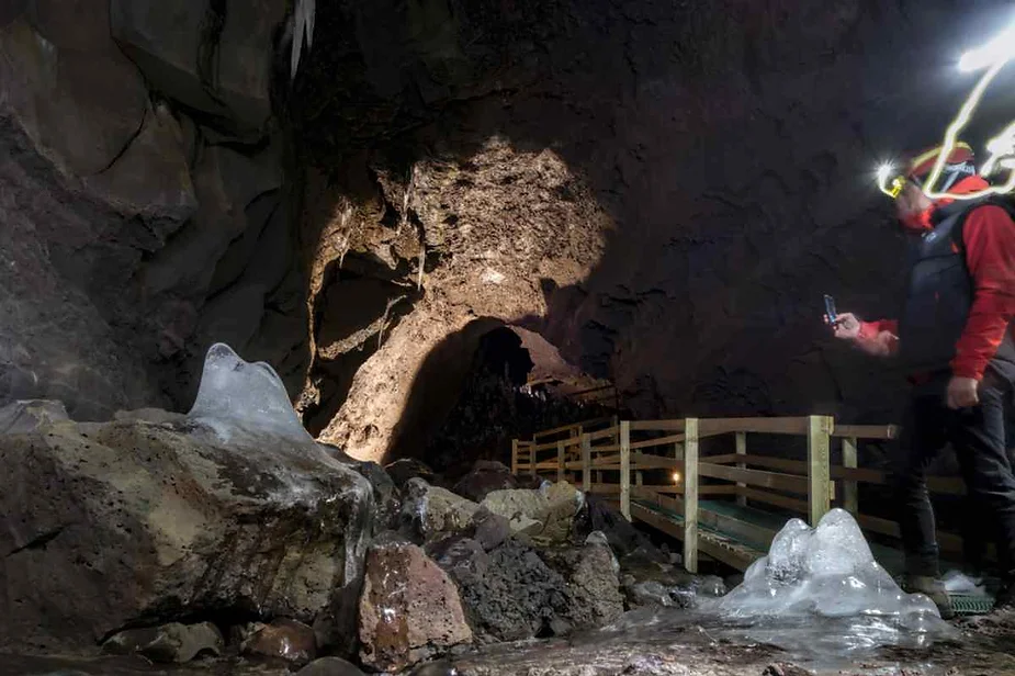 A tourist wearing a headlamp explores Vidgelmir Cave in Iceland, walking along a wooden path next to ice formations and lava rocks, highlighting the cave’s geological wonders.