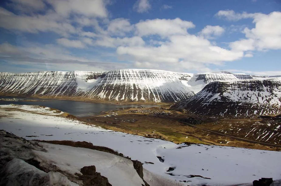 A breathtaking view of snow-capped mountains surrounding a fjord in Iceland, with a small village nestled below under a partly cloudy sky