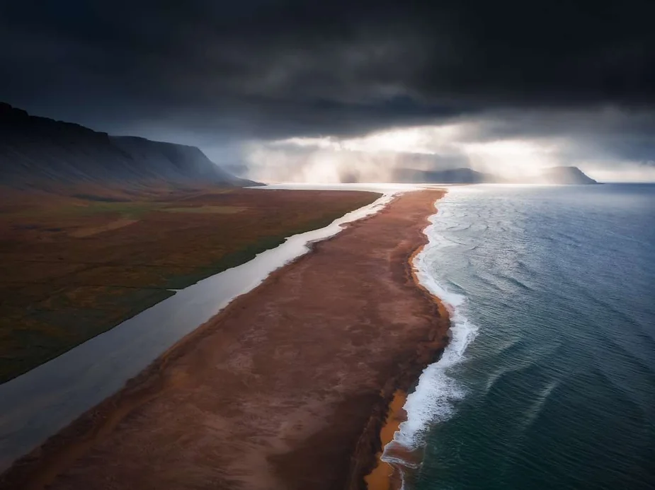 A dramatic aerial view of a narrow strip of land separating the ocean and an inland body of water in Iceland, with dark clouds and sun rays piercing through the sky