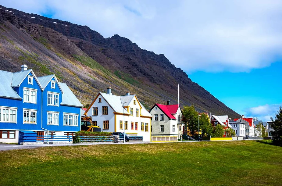 A row of colorful houses in an Icelandic town, with a striking blue house among white and red-roofed buildings, set against a backdrop of rugged mountains and a bright sky