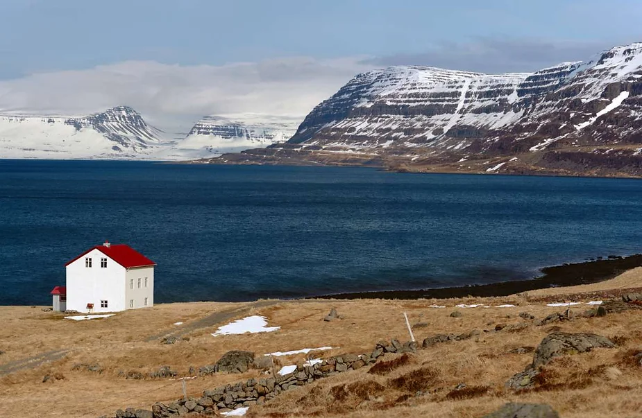 A small white house with a red roof sits near the shore of a fjord in Iceland, surrounded by snow-capped mountains and a blue sea