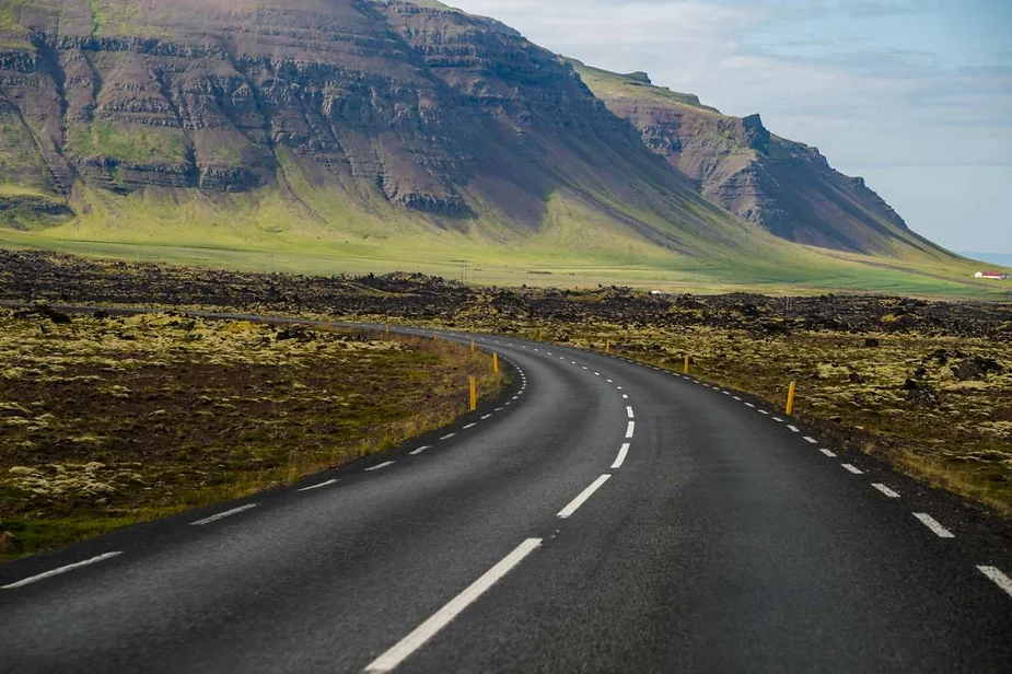 A winding road leading through a rugged, green landscape with towering mountains in the background in Iceland. The road is bordered by yellow markers