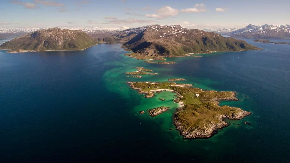 Aerial view of a coastal landscape in Iceland featuring lush green islands and peninsulas surrounded by clear blue waters and mountainous terrain in the background