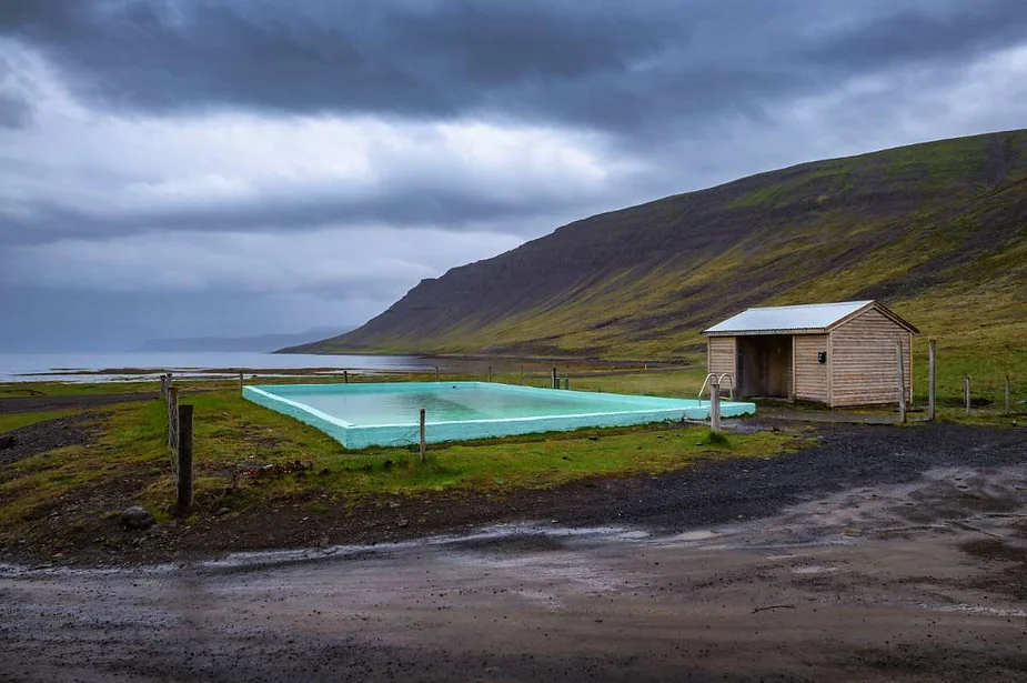 An outdoor hot spring pool in Iceland, surrounded by a rugged landscape with green hills and a cloudy sky, featuring a simple wooden changing shed nearby