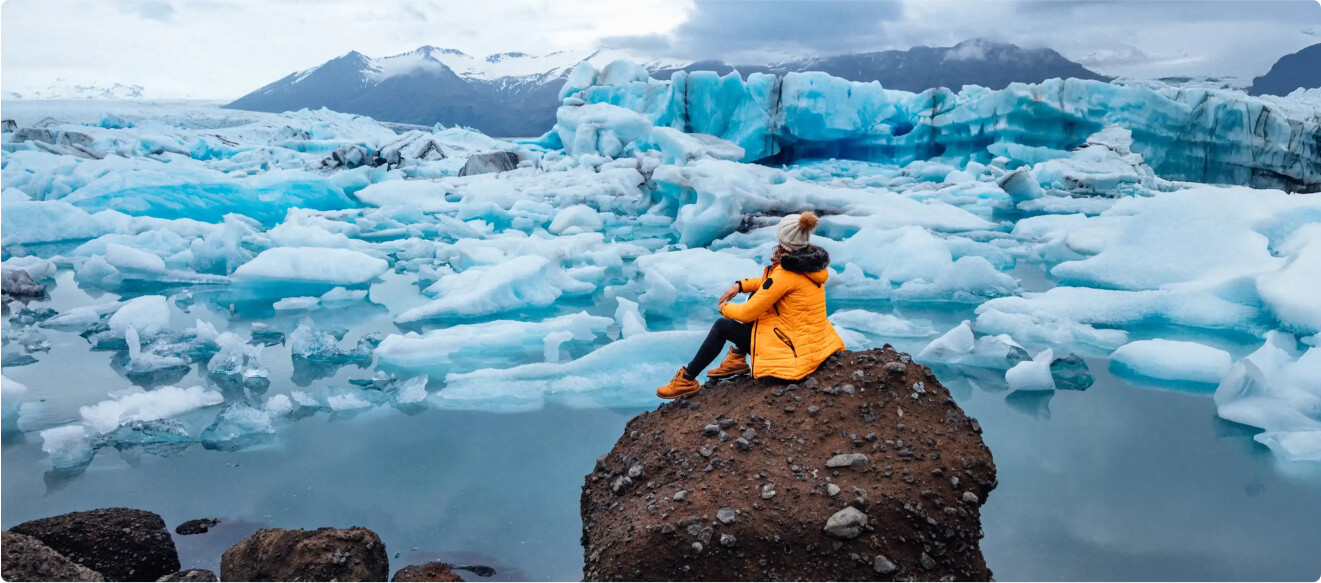 Tourist in yellow winter jacket sitting on a rock in front of a beautiful glacial lake full of icebers