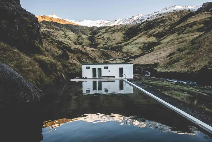 A tranquil geothermal pool in Iceland, surrounded by lush green hills and a small white building, with snow-capped mountains in the background.