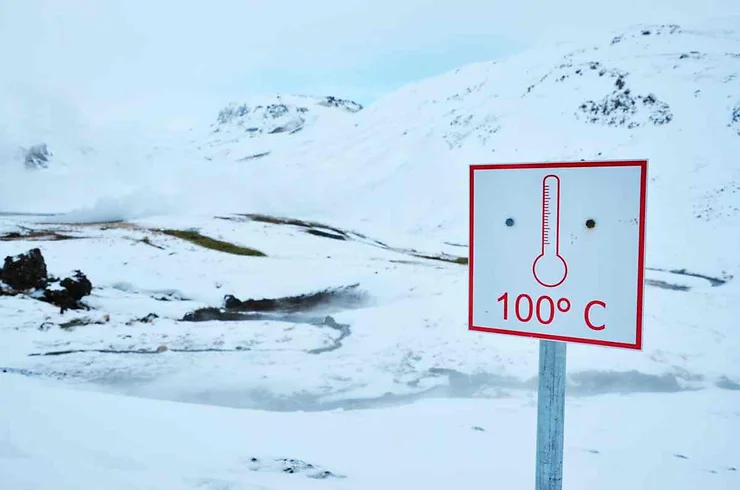 A sign in Iceland indicating a hot spring with a temperature of 100°C, set against a snowy mountainous backdrop.