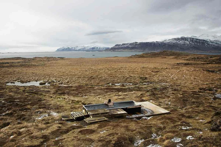 A secluded hot tub in Iceland, situated in a vast, barren landscape with distant snow-capped mountains and a lake under an overcast sky.