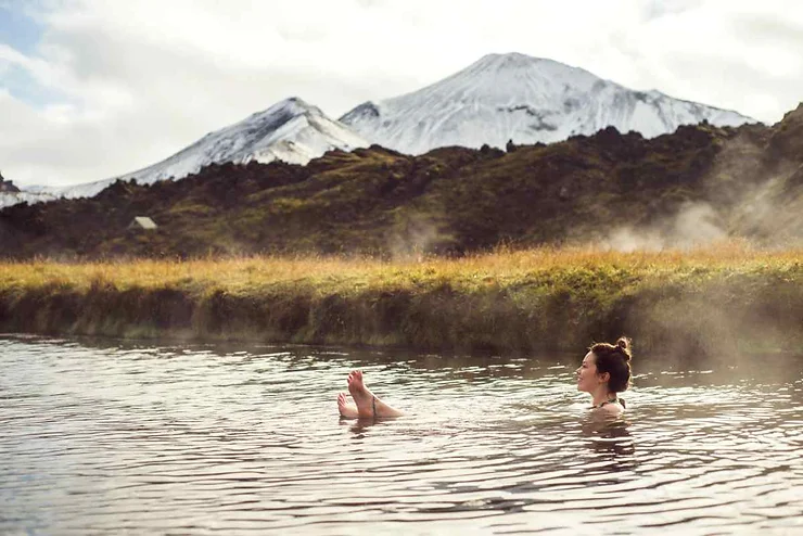 A person relaxing in a warm sulfur spring with steam rising, set against a backdrop of snowy mountains in Iceland.