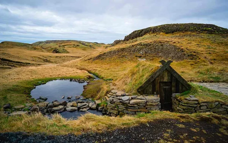 A rustic hot spring in Iceland, surrounded by rocky terrain and gentle hills, featuring a traditional stone structure next to the water.