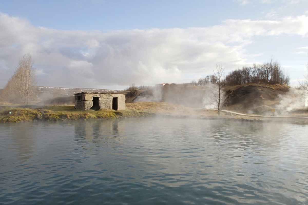 Aerial view of Secret Lagoon with steam rising from the geothermal pool surrounded by green landscape.