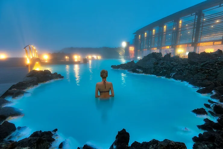 A serene view of a woman relaxing in the warm, mineral-rich waters of the Blue Lagoon in Iceland, surrounded by volcanic rocks and soft, ambient lighting.