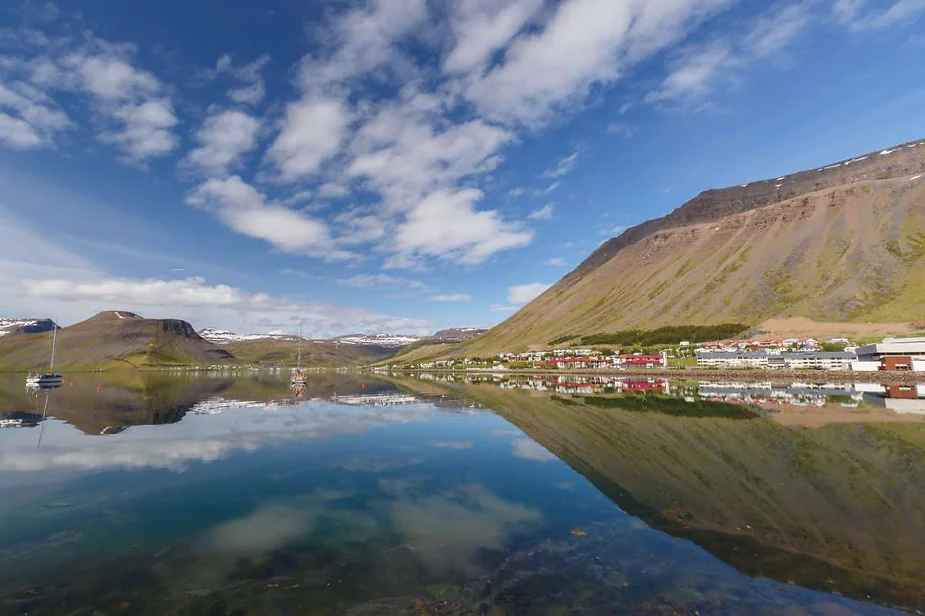 Scenic view of Ísafjörður town in Iceland, reflecting in calm waters surrounded by towering mountains under a bright blue sky.