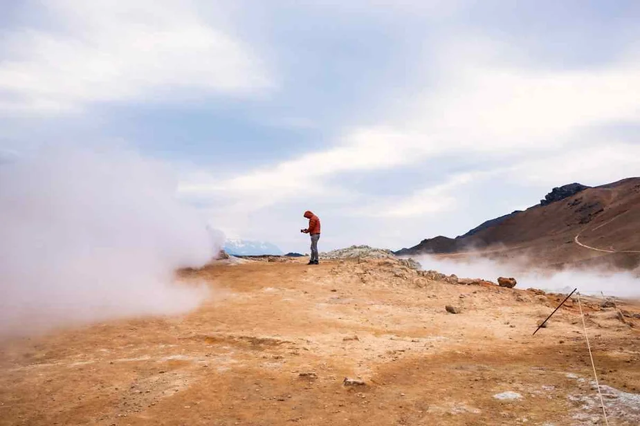 Visitor observing a steaming fumarole at Hverir, Iceland, surrounded by a barren volcanic landscape with dramatic steam clouds rising into the sky.