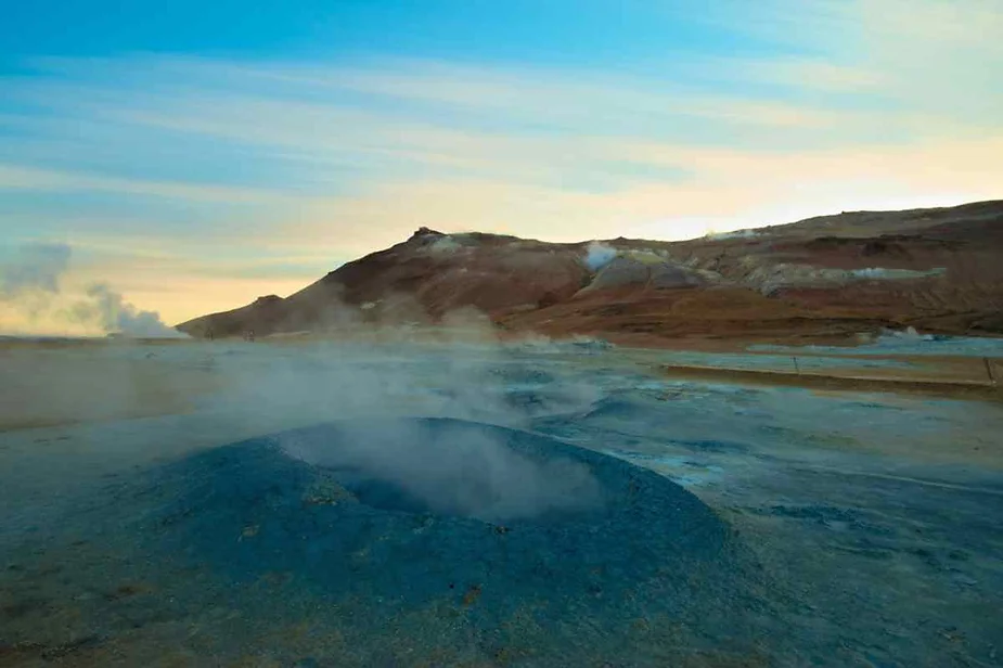 Blue geothermal vent at Hverir, Iceland, emitting steam with vibrant volcanic hills and a dramatic sunrise in the background.