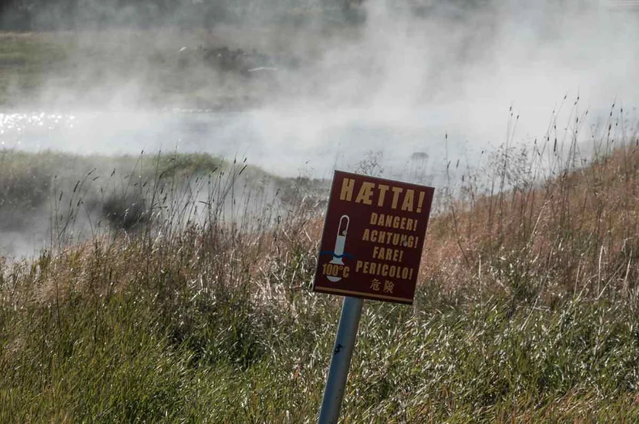 Warning sign at Hverir geothermal area, Iceland, cautioning visitors about 100°C temperatures with steam rising from the surrounding grassy terrain.