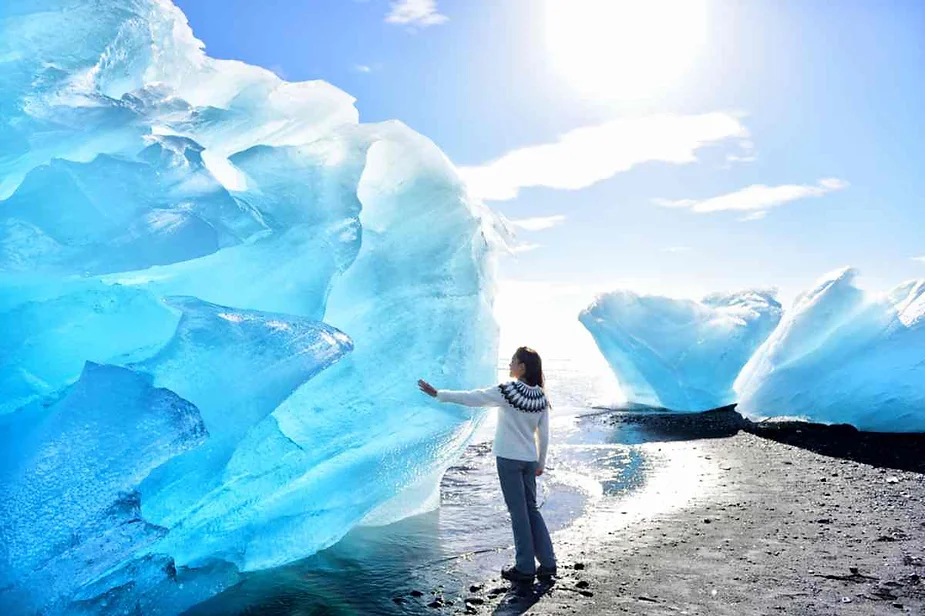 Traveler touching a massive blue iceberg at Diamond Beach, Iceland, with sunlight sparkling on the ice and black sand in the background.