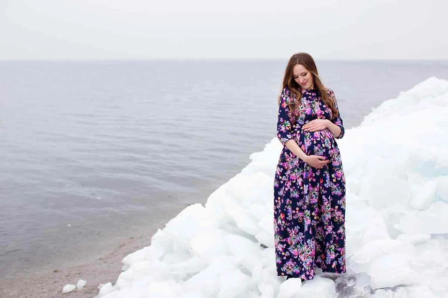 Pregnant woman standing on icy shores in Iceland, wearing a floral dress, embracing her belly while surrounded by serene waters and snow-covered ice.