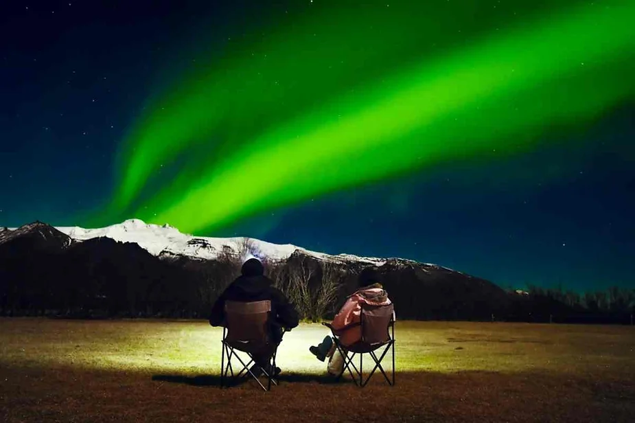 Couple sitting under the Northern Lights in Iceland, enjoying a spectacular aurora display against snow-covered mountains and a starry sky.