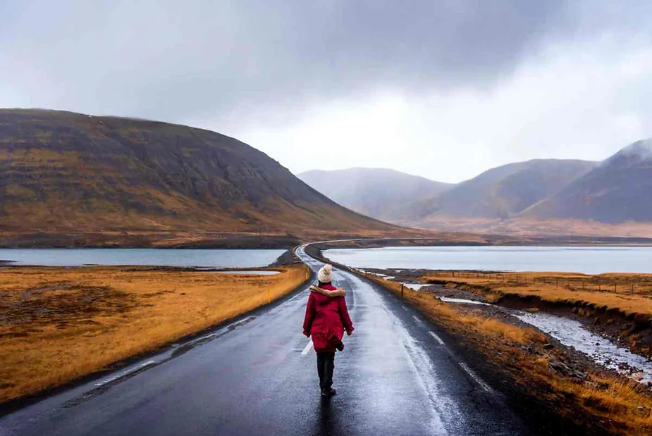 Solo traveler walking on an empty road in Iceland, surrounded by dramatic mountains, lakes, and golden autumn landscapes under a moody sky.