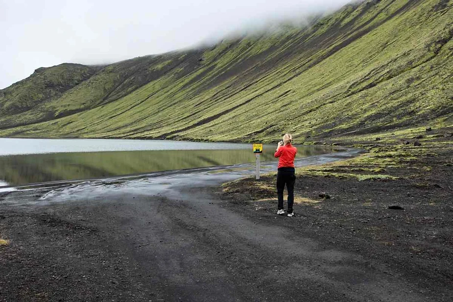 Traveler photographing the serene lake along a challenging dirt track route to Langisjór, surrounded by steep moss-covered volcanic mountains.