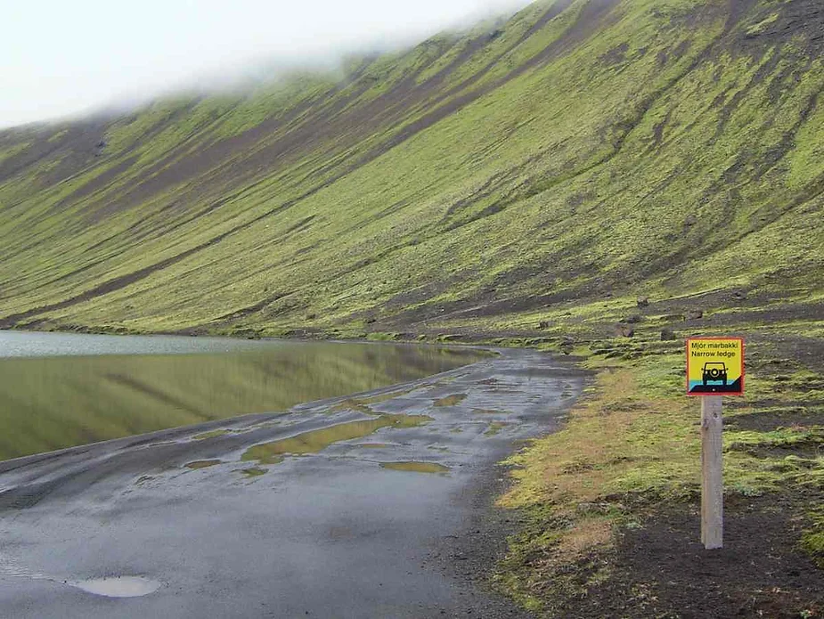 Warning sign marking the narrow ledge along Botnlangalón Route to Langisjór, with mossy volcanic slopes reflected in the calm lake water.