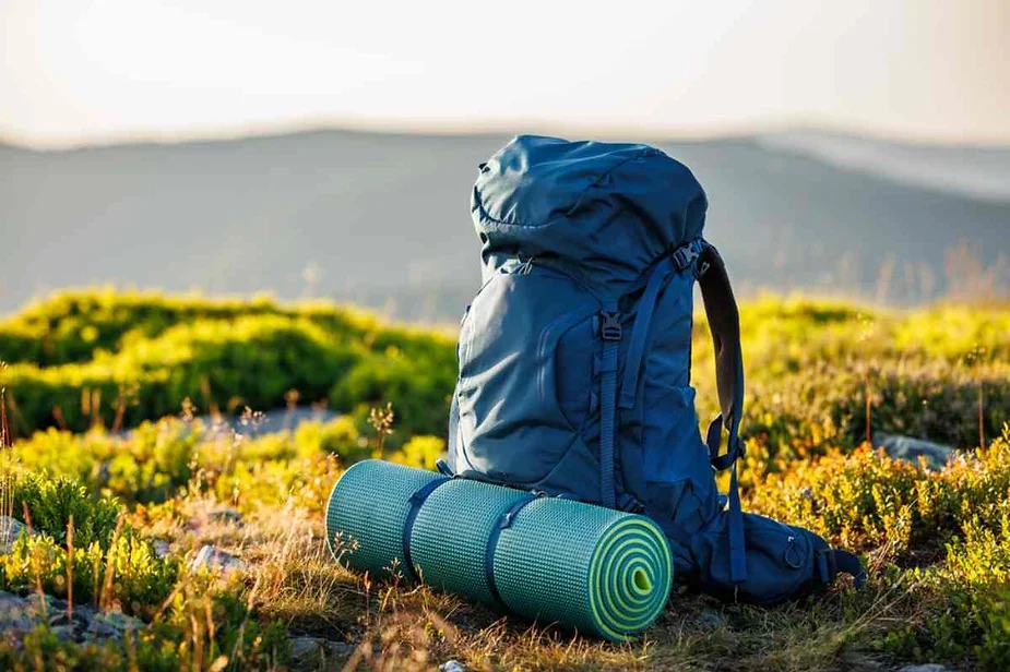 Essential hiking gear including a blue backpack and a rolled-up green sleeping mat, set on a grassy highland with a scenic mountain backdrop.