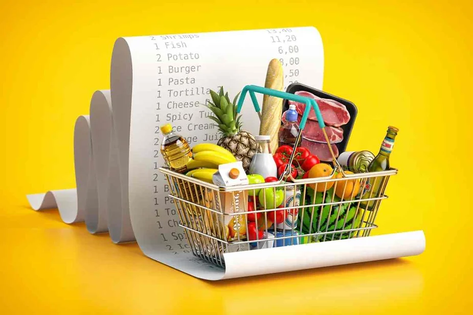 Grocery basket filled with fresh produce, meats, and beverages placed in front of a long grocery receipt on a bright yellow background, illustrating Icelandic grocery pricing.