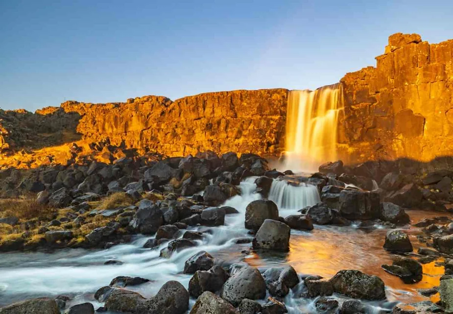 Oxararfoss Waterfall illuminated by golden sunlight during sunset, surrounded by rugged cliffs and a flowing stream in Iceland's Thingvellir National Park.