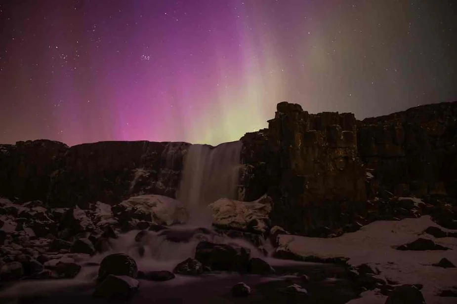 Oxararfoss Waterfall illuminated under vibrant northern lights in Iceland, surrounded by snow-covered rocks and a starry sky.