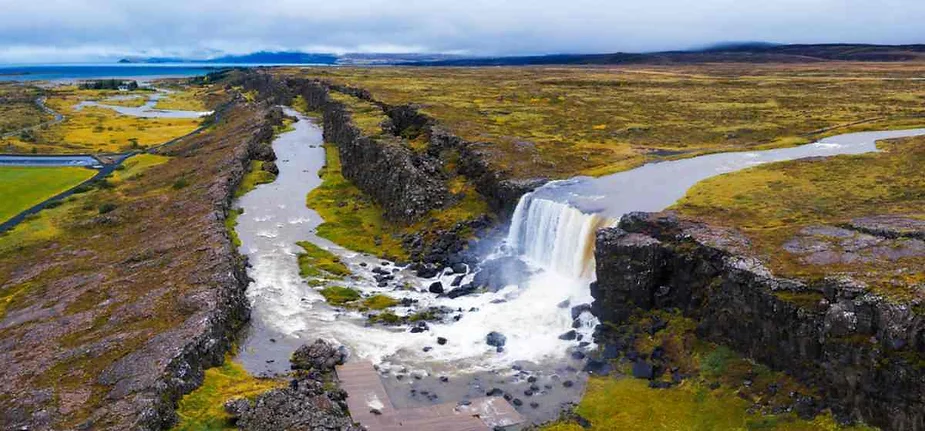 Aerial view of Oxararfoss Waterfall flowing through the Almannagja Gorge with lush moss-covered cliffs and Iceland's vast landscape in the background.