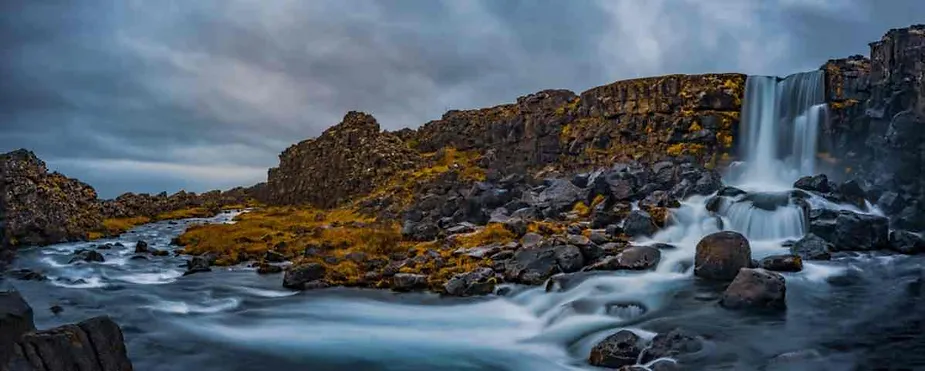 Panoramic view of Oxararfoss Waterfall flowing into a rocky stream, surrounded by moss-covered cliffs under a cloudy Icelandic sky.