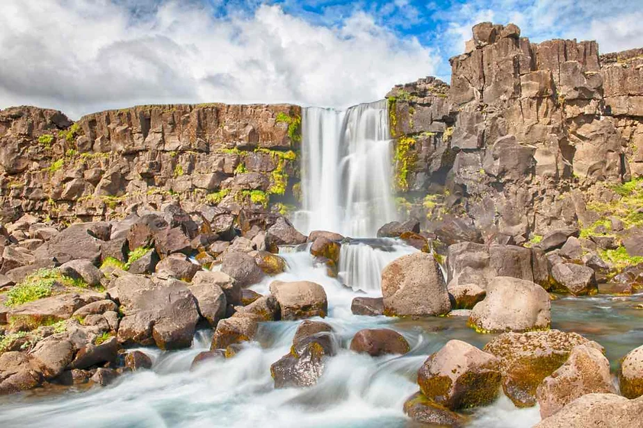 Oxararfoss Waterfall cascading over moss-covered cliffs into a rocky stream under a bright, partly cloudy Icelandic sky.